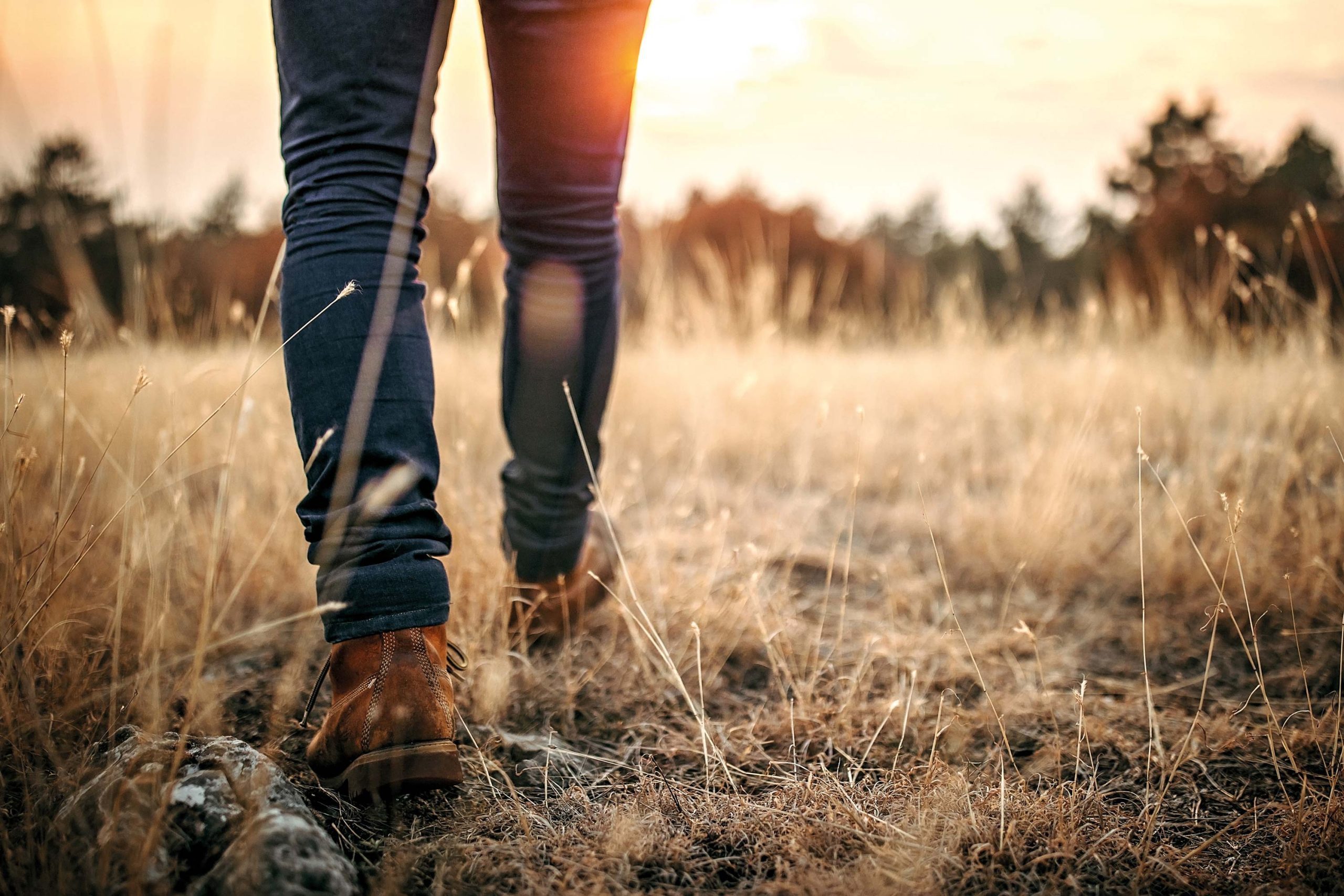 A mans legs and boots walking through a patch of yellow grass at sunset