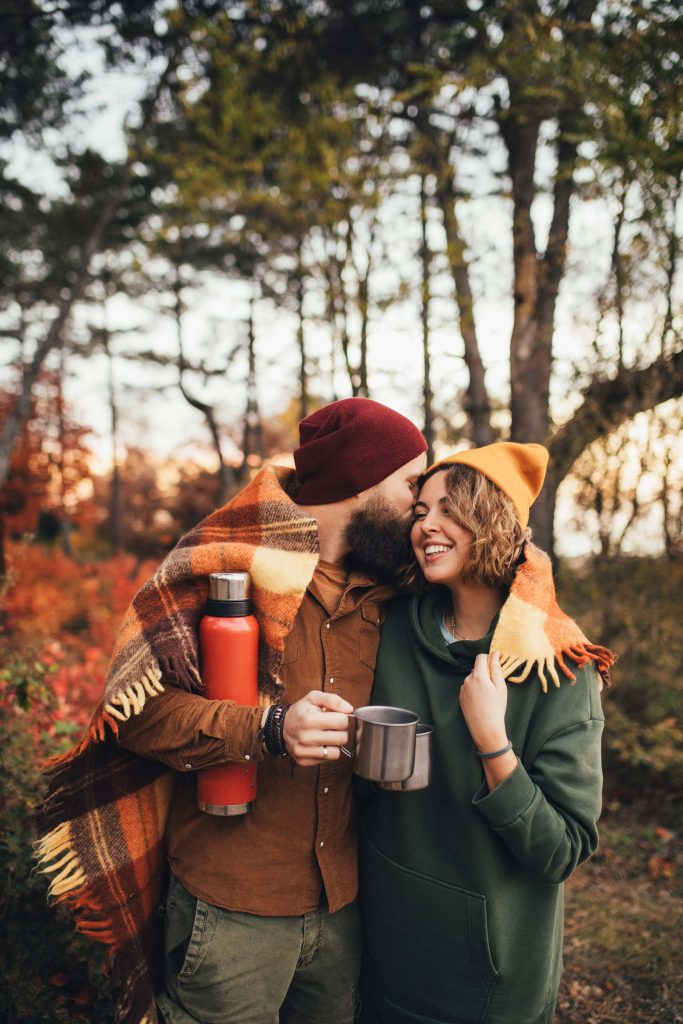 A couple walking through an autumnal forest with a picnic blanket and hot drinks