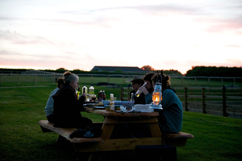 A group of friends enjoying an evening meal on a picnic bench at Loose Reins as the sun sets