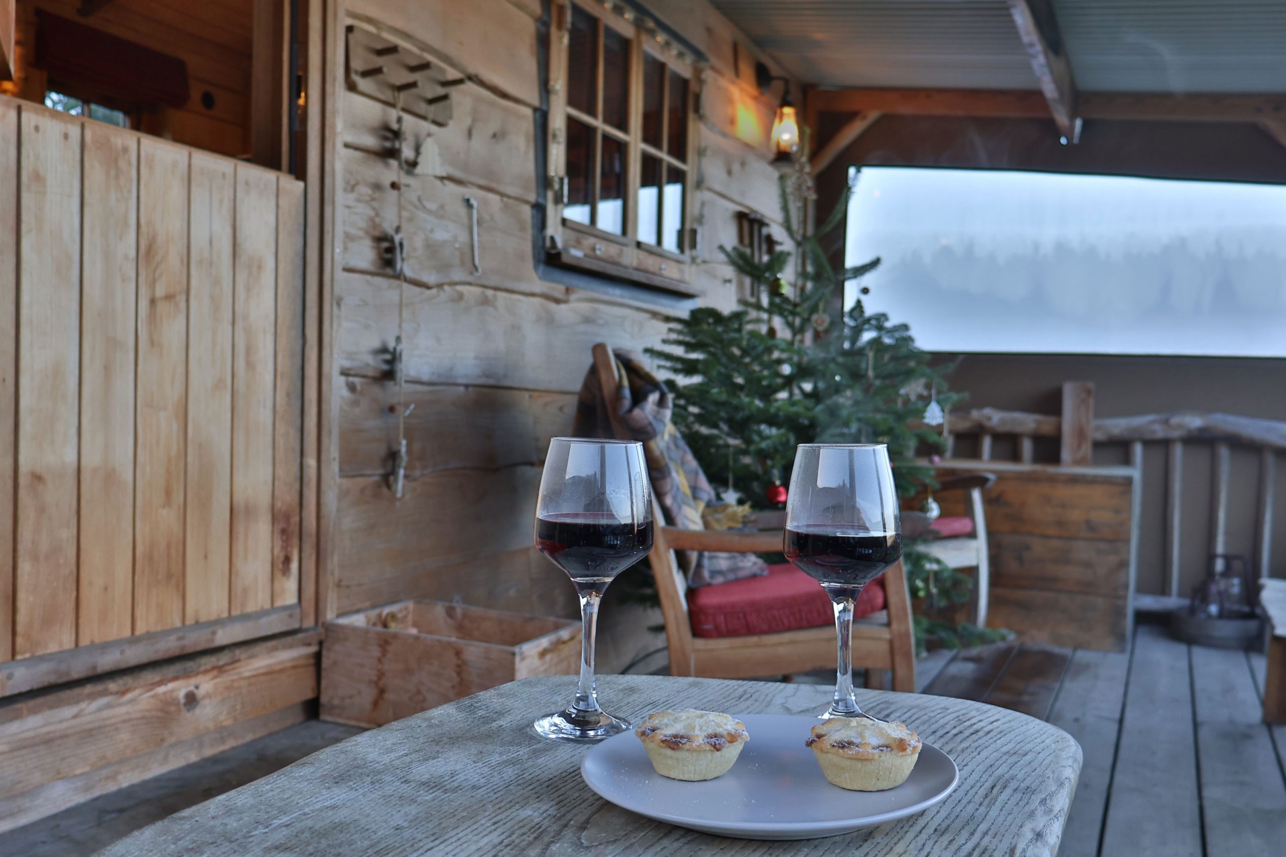 Mince Pies and Mulled Wine on table on porch of rustic glamping cabin which Christmas tree in background