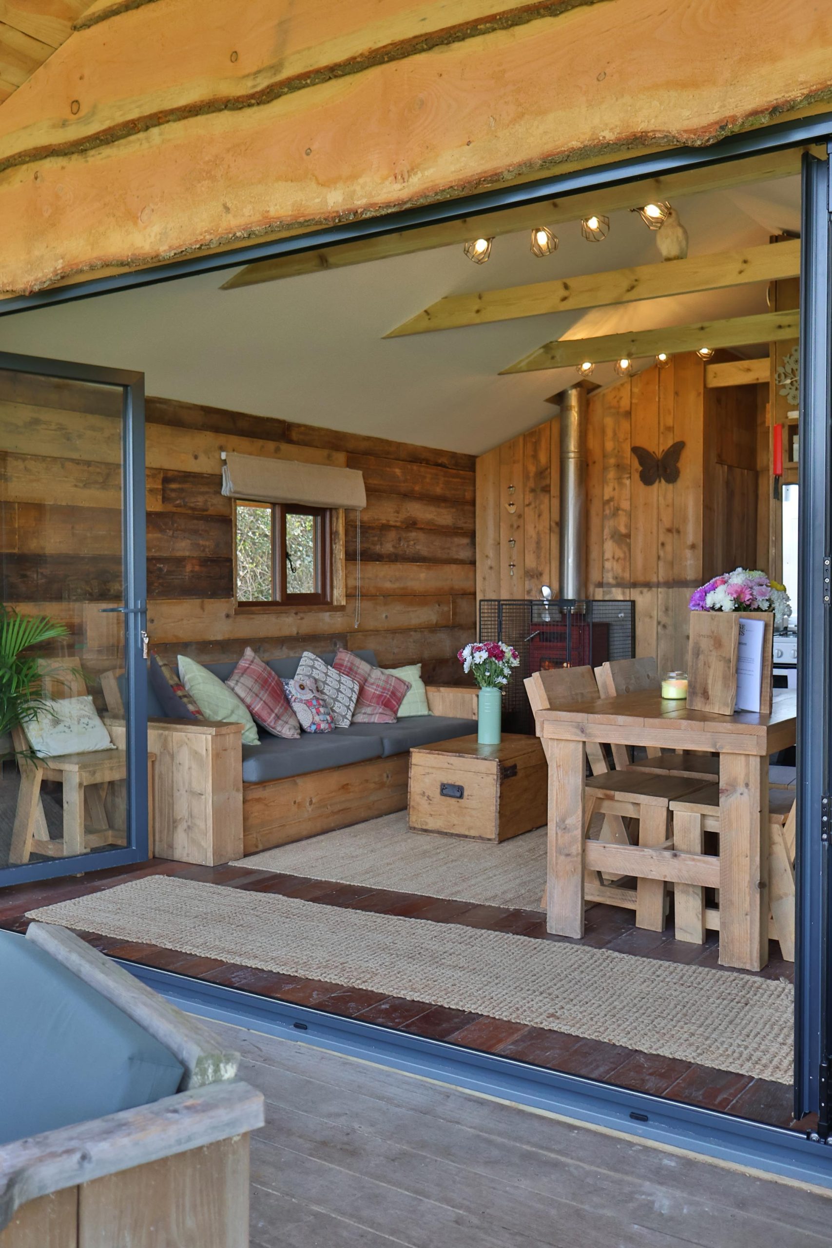 View from the Foresters cabin deck looking in through the bifold doors to the stunning lounge with sofa, wood burner and kitchen table in view.