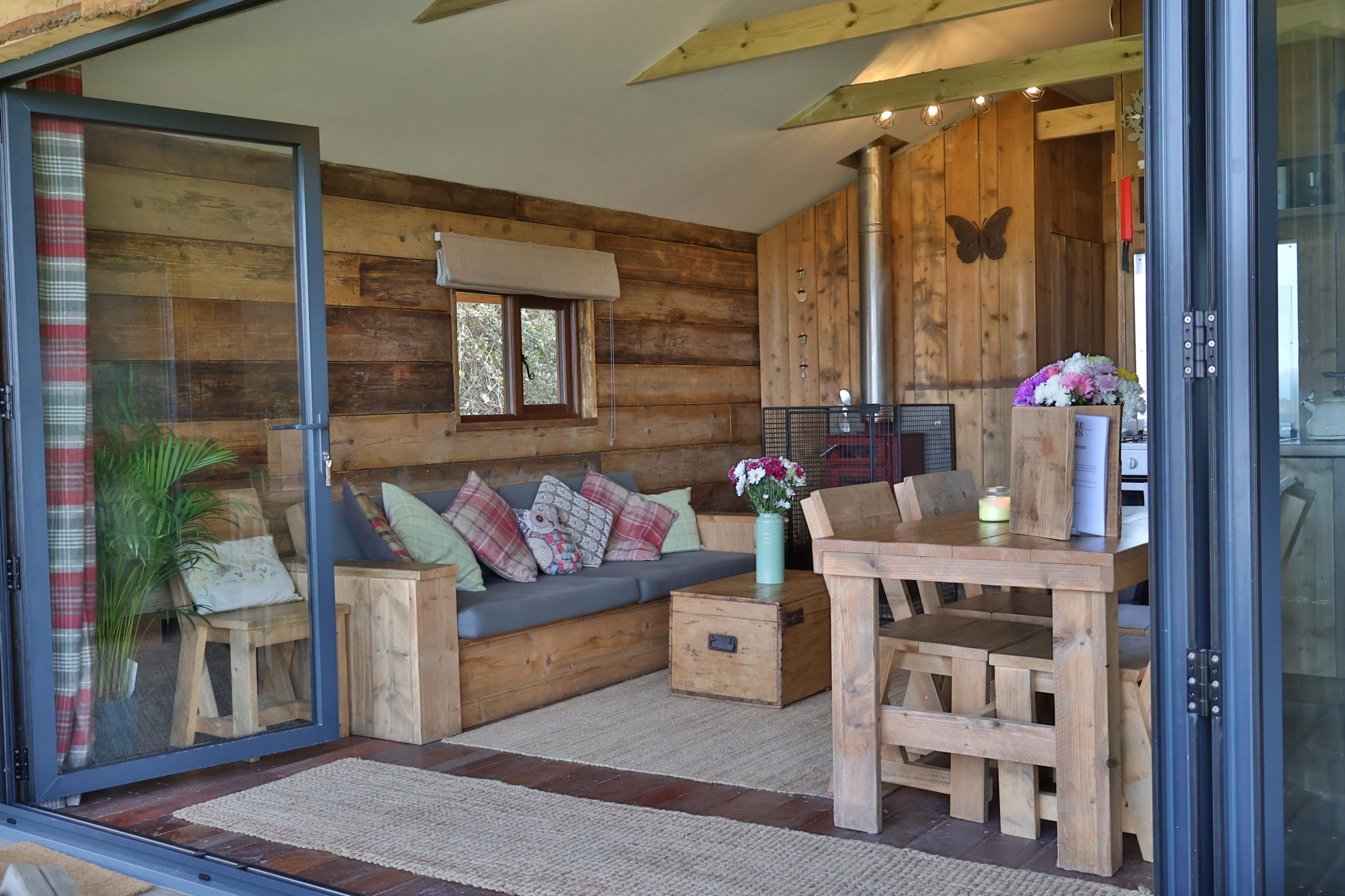 View from the Foresters cabin deck looking in through the bifold doors to the stunning lounge with sofa, wood burner and kitchen table in view.